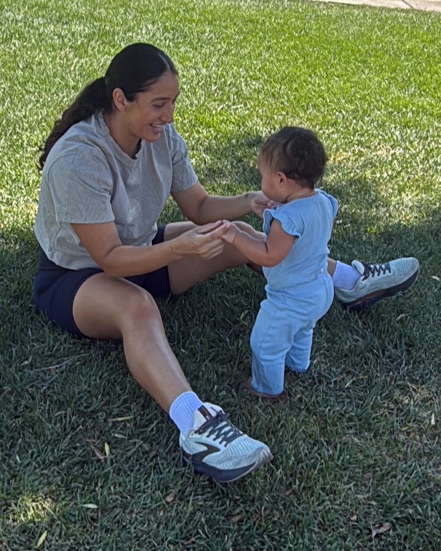 Natalie sitting on the grass at a park, holding her daughter's hands as she stands.